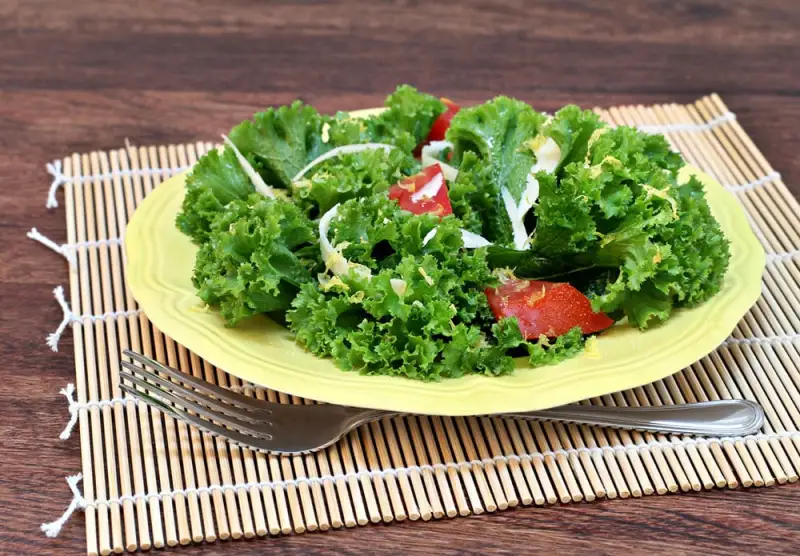 Curly kale salad with tomatoes and cheese on a yellow plate and placemat with fork on a table.