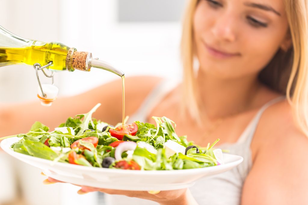 A smiling woman pouring healthy olive oil onto a fresh salad, illustrating why am I craving fatty foods.